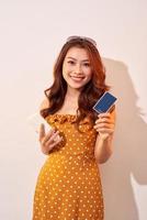imagem de linda jovem isolada sobre fundo bege, mostrando o cartão de crédito plástico enquanto segura o celular foto