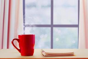 uma caneca vermelha de café quente e notebook ao lado das janelas no inverno e neve caindo foto