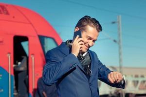 homem feliz falando ao telefone enquanto verifica o tempo no relógio de pulso na estação de trem. foto