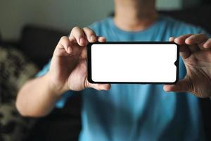 apresentação de tela em branco do smartphone. um jovem está segurando um telefone com uma maquete de tela vazia branca. um homem está vestindo camiseta azul na sala de estar em casa. foto