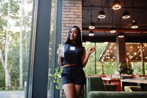 mulher afro-americana feminista na moda usa camiseta preta e shorts, posou no restaurante com copo de limonada. foto