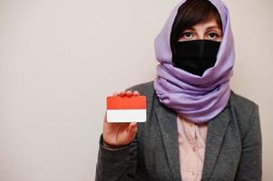 retrato de uma jovem muçulmana vestindo roupa formal, proteger a máscara facial e o lenço de cabeça hijab, segurar o cartão de bandeira da indonésia contra um fundo isolado. conceito de país coronavírus. foto