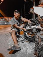 um velho de short curto está se esforçando para trocar o pneu traseiro da motocicleta à noite em sua oficina. foto tirada à noite ao ar livre na vila de luhu, gorontalo, em 21 de agosto de 2022