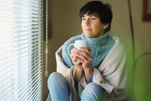 mulher adulta meada tomando café e olhando pela janela em dia de inverno. foto