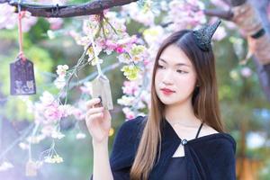 retrato linda mulher asiática que tem boa pele e cabelo comprido em pé segurando uma placa de madeira pendurada em uma árvore veste uma camisa preta enquanto ela sorri alegremente no jardim de flores. foto