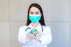 A médica asiática bonita usa máscara facial e segura a bola global nas mãos para salvar o mundo e proteger o conceito mundial. foto