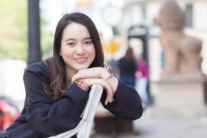 retrato mulher bonita asiática na camisa de manga longa azul escuro senta-se e sorria na cadeira em um parque urbano ao ar livre. foto