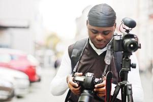 jovem cinegrafista americano africano profissional segurando a câmera profissional com equipamento pro tripé. cinegrafista afro vestindo duraq preto fazendo vídeos. foto