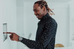 homem afro-americano sorridente usando sistema de casa inteligente moderno, controlador na parede, jovem positivo mudando a temperatura no termostato ou ativando o alarme de segurança no apartamento foto