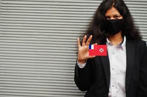 mulher asiática com roupa formal e máscara protetora preta segura a bandeira de wallis e futuna à mão contra um fundo cinza. coronavírus no conceito de país. foto