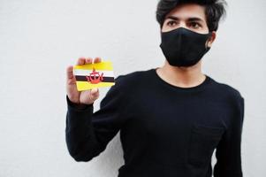 homem asiático usar todo preto com máscara facial segurar a bandeira brunei na mão isolada no fundo branco. conceito de país coronavírus. foto
