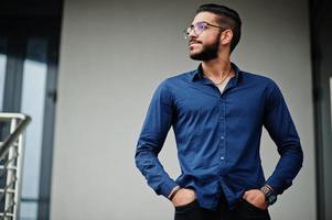 empresário do Oriente Médio usa camisa azul, óculos contra prédio de escritórios. foto