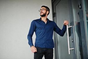 empresário do Oriente Médio usa camisa azul, óculos contra prédio de escritórios. foto
