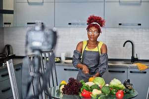 mulher afro-americana filmando sua transmissão de blog sobre comida saudável na cozinha de casa. ela corta um tomate com faca. foto