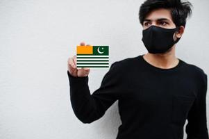 homem asiático usar todo preto com máscara facial segurar a bandeira azad kashmir na mão isolada no fundo branco. conceito de país coronavírus. foto