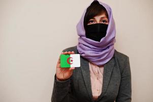 retrato de uma jovem muçulmana vestindo roupa formal, proteger a máscara facial e o lenço de cabeça hijab, segurar o cartão de bandeira da argélia contra um fundo isolado. conceito de país coronavírus. foto