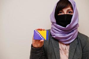 retrato de uma jovem muçulmana vestindo roupa formal, proteger a máscara facial e o lenço de cabeça hijab, segurar o cartão de bandeira da bósnia e herzegovina contra um fundo isolado. conceito de país coronavírus. foto