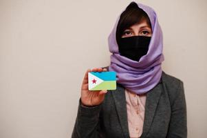 retrato de jovem muçulmana vestindo roupa formal, proteger máscara facial e lenço de cabeça hijab, segure o cartão de bandeira do djibuti contra fundo isolado. conceito de país coronavírus. foto