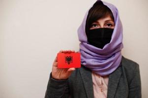 retrato de jovem muçulmana vestindo roupa formal, proteger máscara facial e lenço de cabeça hijab, segurar o cartão de bandeira da albânia contra fundo isolado. conceito de país coronavírus. foto