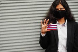 mulher asiática com roupa formal e máscara protetora preta segura a bandeira do havaí à mão contra um fundo cinza. coronavírus no conceito de país. foto