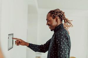 homem afro-americano sorridente usando sistema de casa inteligente moderno, controlador na parede, jovem positivo mudando a temperatura no termostato ou ativando o alarme de segurança no apartamento foto