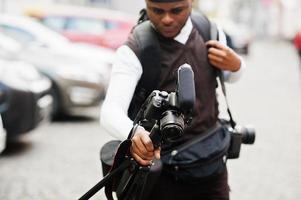 jovem cinegrafista americano africano profissional segurando a câmera profissional com equipamento pro tripé. cinegrafista afro vestindo duraq preto fazendo vídeos. foto