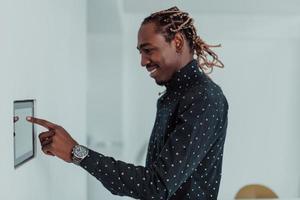 homem afro-americano sorridente usando sistema de casa inteligente moderno, controlador na parede, jovem positivo mudando a temperatura no termostato ou ativando o alarme de segurança no apartamento foto