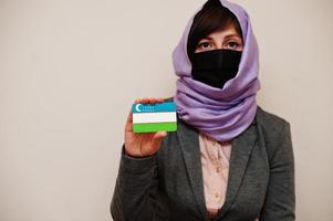 retrato de jovem muçulmana vestindo roupa formal, proteger máscara facial e lenço de cabeça hijab, segure o cartão de bandeira do uzbequistão contra fundo isolado. conceito de país coronavírus. foto