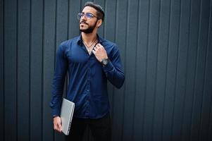 empresário do Oriente Médio usa camisa azul, óculos com laptop contra parede de aço. foto