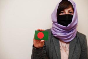 retrato de jovem muçulmana vestindo roupa formal, proteger máscara facial e lenço de cabeça hijab, segure o cartão de bandeira de bangladesh contra fundo isolado. conceito de país coronavírus. foto