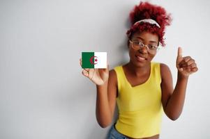 mulher africana com cabelo afro, use camiseta amarela e óculos, segure a bandeira da argélia isolada no fundo branco, mostre o polegar para cima. foto