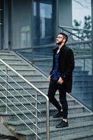 empresário do oriente médio usa casaco preto e camisa azul, óculos contra prédio de escritórios, fica nas escadas.