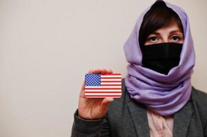 retrato de uma jovem muçulmana vestindo roupa formal, proteger a máscara facial e o lenço de cabeça hijab, segurar o cartão de bandeira dos eua contra um fundo isolado. conceito de país coronavírus. foto