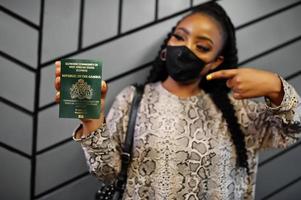 mulher africana usando máscara facial preta mostra passaporte da gâmbia na mão. coronavírus no país africano, fechamento e quarentena de fronteiras, conceito de surto de vírus. foto