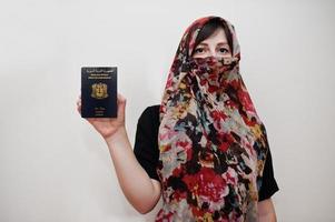 jovem muçulmana árabe em roupas hijab segura passaporte da república árabe síria no fundo da parede branca, retrato de estúdio. foto