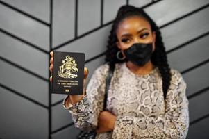 mulher afro-americana usando máscara facial preta mostra passaporte das bahamas na mão. coronavírus no país da américa, fechamento de fronteiras e quarentena, conceito de surto de vírus. foto