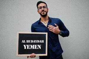homem árabe usa camisa azul e óculos segura placa com inscrição al hudayah yemen. maiores cidades no conceito do mundo islâmico. foto