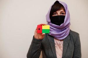 retrato de uma jovem muçulmana vestindo roupa formal, proteger a máscara facial e o lenço de cabeça hijab, segurar o cartão de bandeira da Guiné-bissau contra um fundo isolado. conceito de país coronavírus. foto