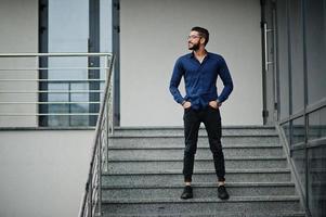 empresário do Oriente Médio usa camisa azul, óculos contra prédio de escritórios. foto