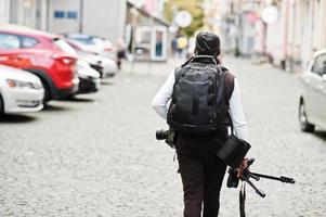 jovem cinegrafista americano africano profissional segurando a câmera profissional com equipamento pro tripé. cinegrafista afro vestindo duraq preto fazendo vídeos. foto