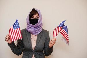 retrato de jovem muçulmana vestindo roupa formal, proteger máscara facial e lenço de cabeça hijab, segurar bandeiras dos eua nas mãos contra fundo isolado. conceito de país coronavírus. foto