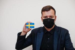 homem europeu usa preto formal e protege a máscara facial, segura o cartão da bandeira da suécia isolado no fundo branco. conceito de país covid coronavírus da europa. foto