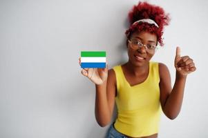 mulher africana com cabelo afro, use camiseta amarela e óculos, segure a bandeira de serra leoa isolada no fundo branco, mostre o polegar para cima. foto