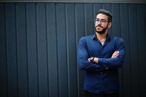 empresário do Oriente Médio usa camisa azul, óculos, contra a parede de aço.