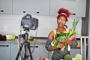 mulher afro-americana filmando sua transmissão de blog sobre comida saudável na cozinha de casa. ela segura legumes nas mãos. foto