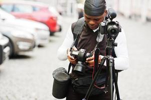 jovem cinegrafista americano africano profissional segurando a câmera profissional com equipamento pro tripé. cinegrafista afro vestindo duraq preto fazendo vídeos. foto