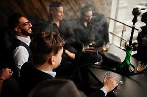 grupo de gângsteres de homem bem vestido retrô bonito passa o tempo no clube, fumando narguilé. festa de máfia de solteiro masculino multiétnica no restaurante. foto