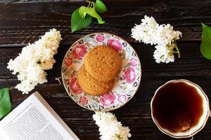 romântica natureza morta com flores lilás. xícara de chá preto, livro aberto e dois biscoitos de aveia em fundo escuro de madeira. foto