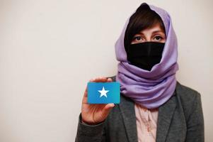 retrato de uma jovem muçulmana vestindo roupa formal, proteger a máscara facial e o lenço de cabeça hijab, segurar o cartão da bandeira da Somália contra um fundo isolado. conceito de país coronavírus. foto