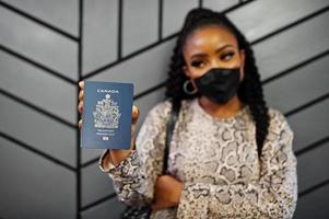 mulher afro-americana usando máscara facial preta mostra passaporte do canadá na mão. coronavírus no país da américa, fechamento de fronteiras e quarentena, conceito de surto de vírus. foto